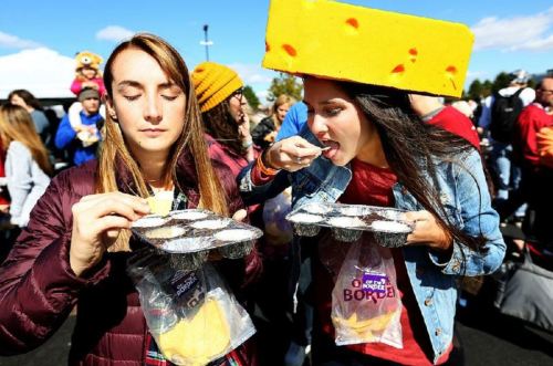 Two women at a festival, one wearing a large cheese hat, both enjoying cheese samples from containers.