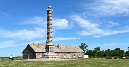 A tall stone monument stands beside a historic building under a blue sky with scattered clouds. Green grass surrounds the area.