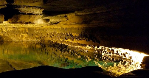 A dimly lit cave with a still water pool reflecting the rocky walls and ceiling, creating a serene atmosphere.