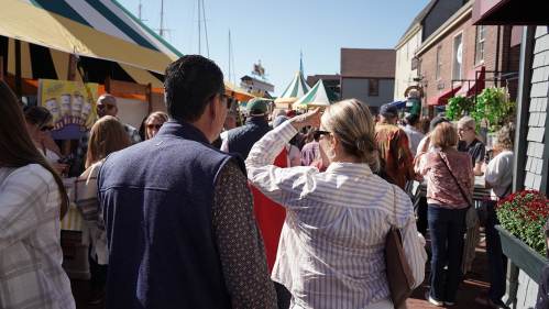 A busy outdoor market scene with people walking among colorful tents and stalls under a clear blue sky.