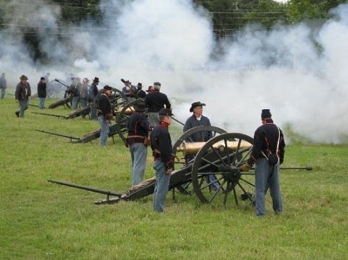 Civil War reenactors operate cannons in a smoky field, dressed in period uniforms.