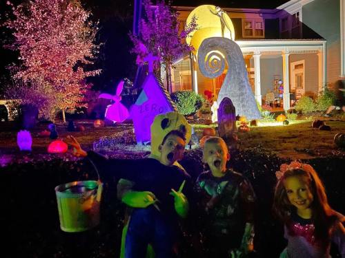 Two children in costumes pose in a spooky, brightly lit yard decorated for Halloween, with pumpkins and eerie figures.