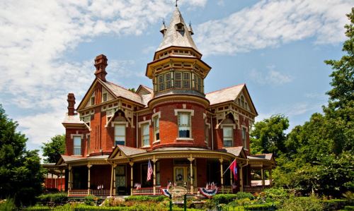 A large, ornate Victorian-style house with a turret, surrounded by greenery and flags, under a blue sky.