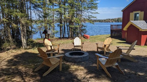A lakeside fire pit area with wooden chairs surrounded by trees and a red cabin in the background.