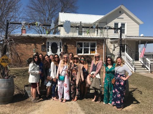 A group of women in floral dresses poses outside a house on a sunny day, with a sign that says "Wine Ahead."
