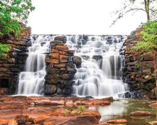 A cascading waterfall flows over rocky formations, surrounded by lush greenery and a calm pool at the base.