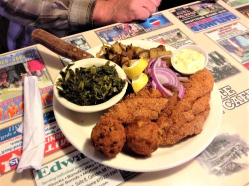 A plate of fried fish, hushpuppies, collard greens, potatoes, and lemon slices on a table covered with menus.