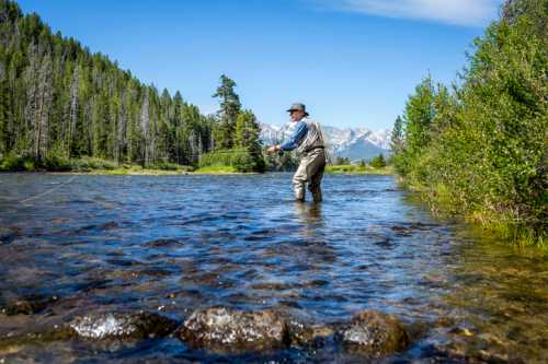 A person fly fishing in a clear river surrounded by lush trees and mountains under a blue sky.