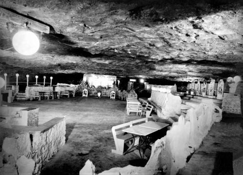A dimly lit underground cavern with stone walls, featuring tables, chairs, and illuminated by hanging lights.
