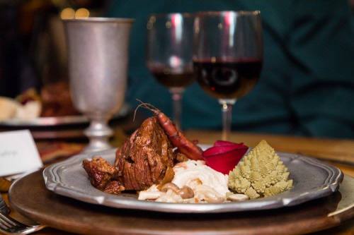 A gourmet plate featuring meat, shrimp, creamy sauce, and a decorative rose, with wine glasses in the background.