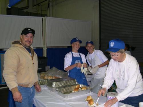 A group of four people in a kitchen setting, preparing food and serving at a table with trays of buns.