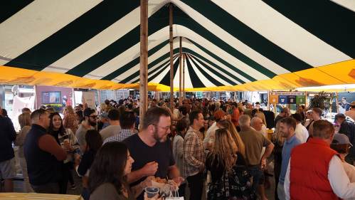 A large crowd of people socializing under a striped tent at an outdoor event.