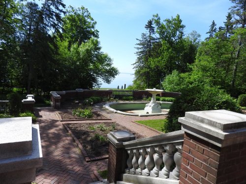 A serene garden view with a fountain, surrounded by lush greenery and a glimpse of water in the distance.