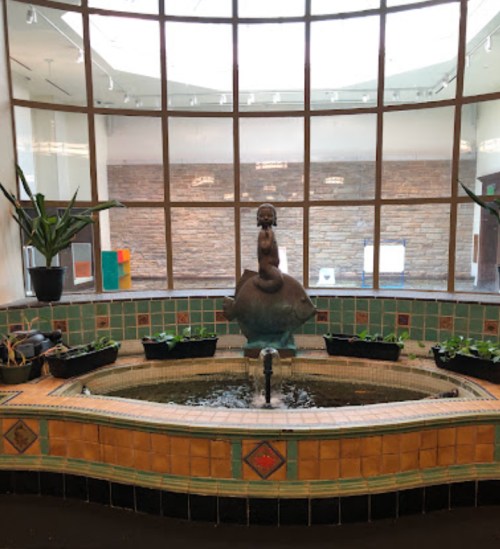 A decorative indoor fountain with a statue, surrounded by plants and colorful tiles, under a large glass ceiling.