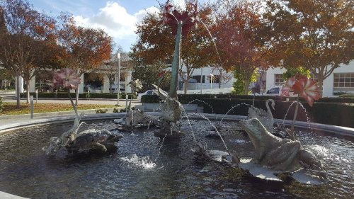 A decorative fountain featuring sculptures of frogs and flowers, surrounded by trees and a clear blue sky.