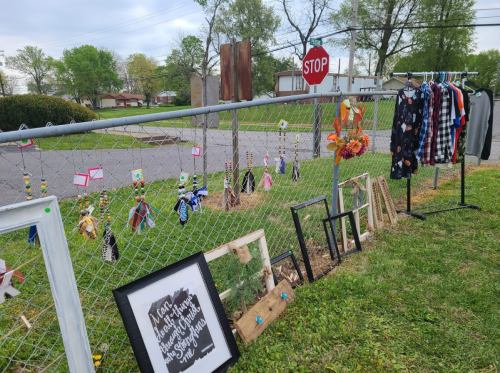 A fence displays handmade crafts, including colorful decorations, frames, and clothing, with a stop sign in the background.