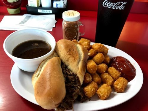 A plate with two sandwiches, tater tots, a bowl of sauce, and a drink on a red table.