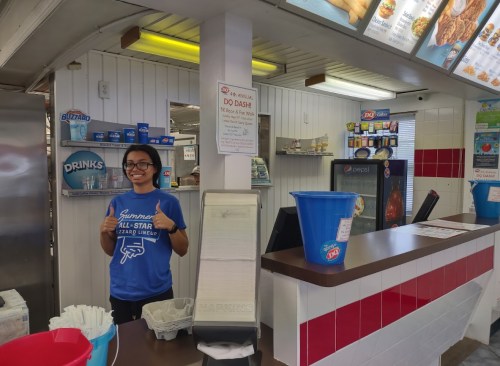 A smiling employee stands behind the counter of an ice cream shop, giving a thumbs up in a blue shirt.
