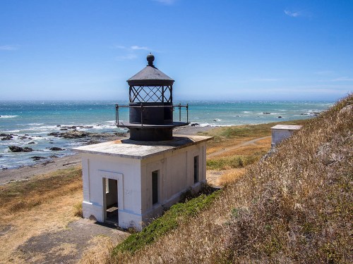 A coastal lighthouse stands near the ocean, surrounded by dry grass and rocky shoreline under a clear blue sky.