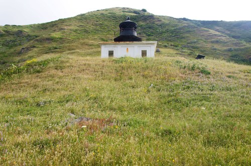 A white lighthouse sits atop a grassy hill, surrounded by wildflowers and rolling green terrain under a cloudy sky.