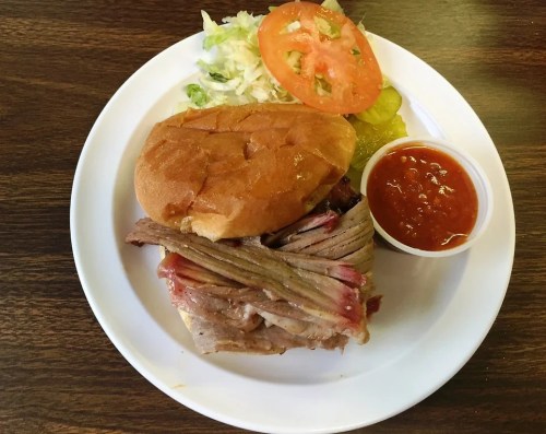 A sandwich with stacked meat on a bun, served with a side of salad, tomato, pickles, and a small cup of sauce.