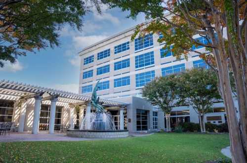 A modern building with large windows, surrounded by trees and a fountain in a landscaped area.