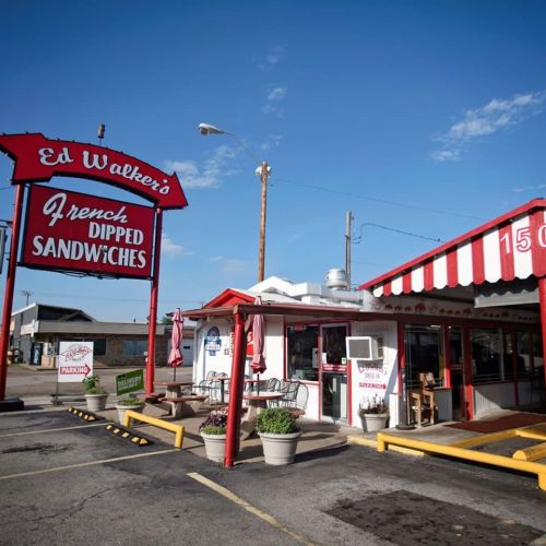 A retro diner with a red and white awning, featuring a sign for "Ed Walker's French Dipped Sandwiches" and outdoor seating.