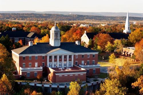Aerial view of a brick building surrounded by colorful autumn trees and distant hills.