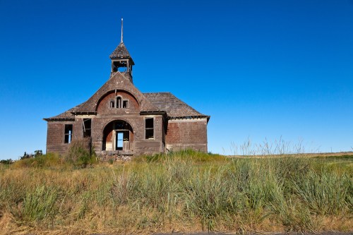 Abandoned wooden schoolhouse with a bell tower, surrounded by tall grass under a clear blue sky.
