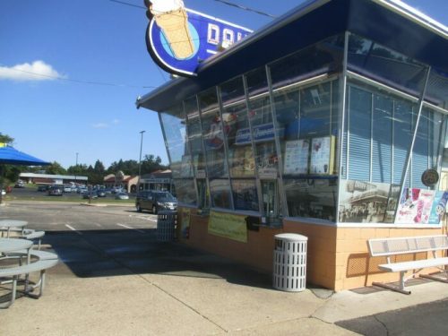 A bright ice cream shop with large windows, outdoor seating, and a blue awning on a sunny day.