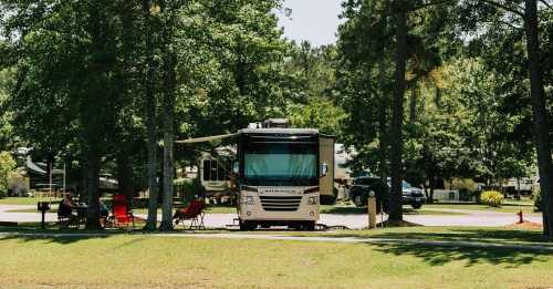 A parked RV in a shaded campsite with chairs set up nearby, surrounded by trees and other RVs in the background.