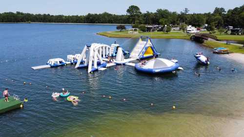 A vibrant water park with inflatable structures on a lake, people swimming, and boats nearby under a clear blue sky.