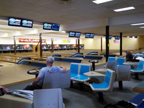 A nearly empty bowling alley with blue seating, bowling lanes, and screens displaying scores. Two people are seated.