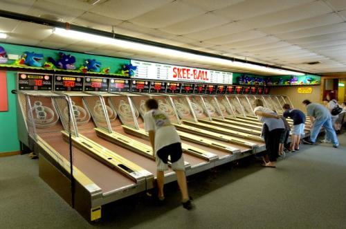 A lively arcade with people playing skee-ball at multiple lanes, colorful decorations in the background.
