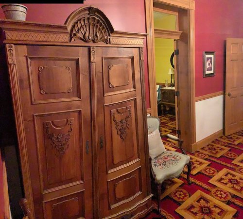 A vintage wooden wardrobe stands against a red wall, with a decorative chair nearby and a doorway leading to another room.