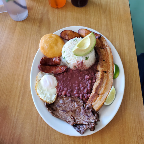 A plate of food featuring rice, beans, fried plantains, steak, sausage, an egg, and a slice of avocado.