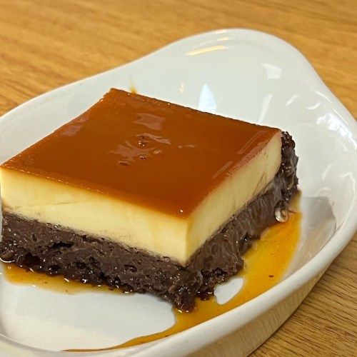 A slice of layered dessert with a smooth caramel top, creamy custard, and a chocolate base on a white plate.