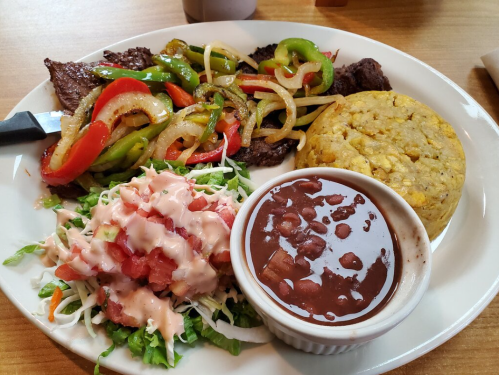 A plate of grilled steak with sautéed peppers and onions, cornbread, salad with dressing, and a side of beans.