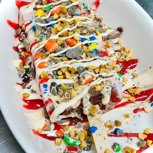 A colorful dessert with a popsicle topped with nuts, candy pieces, and drizzled with white and red sauces.