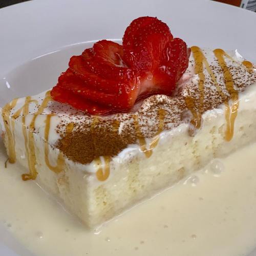 A slice of creamy dessert topped with strawberries, drizzled with caramel and dusted with cocoa powder on a white plate.