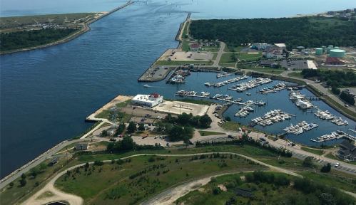 Aerial view of a marina with boats, parking, and a waterway leading to the ocean, surrounded by greenery.