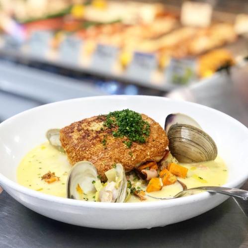 A bowl of creamy seafood chowder topped with a crispy fish fillet and garnished with chives, surrounded by clams and vegetables.
