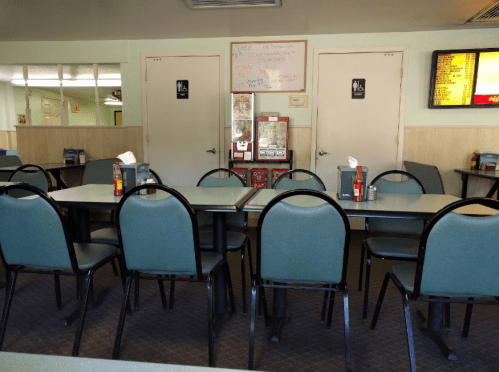 Empty restaurant interior with green chairs, tables, and two closed doors. A menu board is visible in the background.