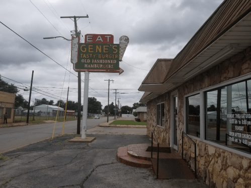 Sign for Gene's Tasty Burger, featuring "Eat Gene's" and "Old Fashioned Hamburgers," outside a stone building on a cloudy day.
