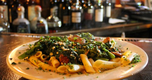 A plate of pasta with greens and herbs, garnished with parsley, set against a blurred background of bottles.