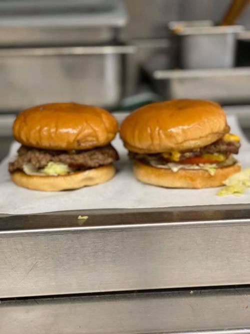 Two burgers on a metal surface, each with a beef patty, cheese, pickles, and sauces, on toasted buns.