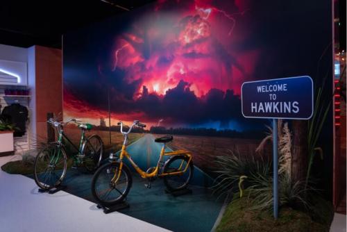 A vibrant backdrop of stormy skies with two bicycles and a "Welcome to Hawkins" sign in a themed display.