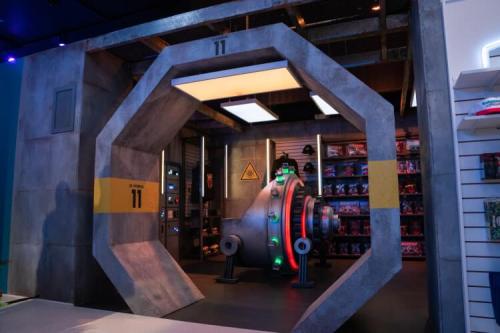 A large, futuristic vault door with green and red lights, set in a concrete wall with shelves of items behind it.