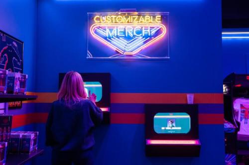 A person interacts with a touchscreen in a vibrant store, under a neon sign that reads "Customizable Merch."