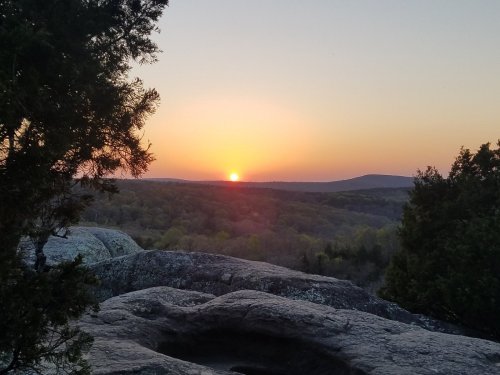 Sunset over a forested landscape, viewed from rocky terrain, with warm hues illuminating the sky.